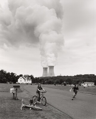 A black-and-white photograph of three White children, two with bikes and one running, on a rural street as two nuclear power plants billow steam in the background.