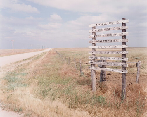 A color photograph of a faded signpost on a desolate country road lined with a barbed wire fence and telephone poles.