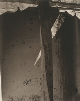 A black-and-white photograph of a wood post, decorative corbels, and a whitewashed wall in shadow.