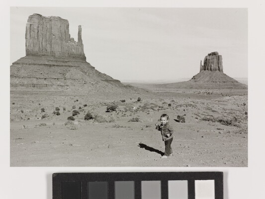 A black-and-white photograph of a toddler in front of tall buttes rising from the desert ground.