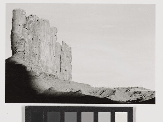 A black-and-white photograph of tall desert rock formation with a shadow encroaching from the bottom.
