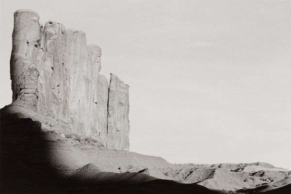 A black-and-white photograph of tall desert rock formation with a shadow encroaching from the bottom.
