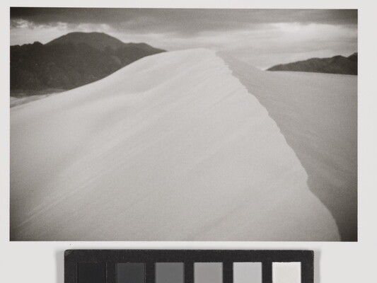 A black-and-white landscape photograph of the ridge of a sand dune against dark mountains and cloudy sky.
