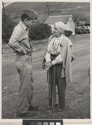 A black-and-white photograph of a White man and a White woman talking to each other on a gravel road with a wood shed in the background.