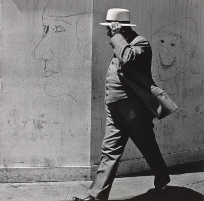 A black-and-white photograph of a man in a three-piece suit and hat walking past a wall covered with graffiti.