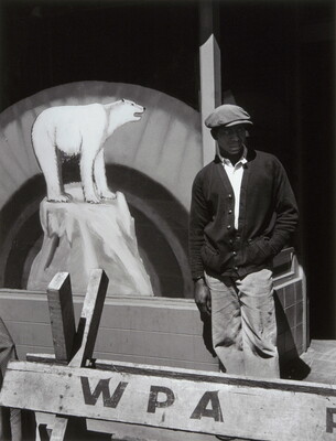 A black-and-white photograph of a Black man in a flat cap and dark cardigan behind a wooden barricade that says "WPA" and in front of a sign with a painting of a polar bear.