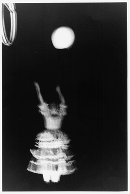 A black-and-white photograph of a person in motion wearing a white dress with a ruffled skirt, arms straight up as if reaching for the circular object above them.