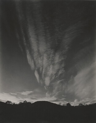 A black-and-white photograph of hills and trees silhouetted against a sky with thin, vertical clouds.