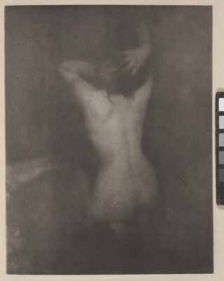 A dark, soft-focus, black-and-white photograph of a nude White woman kneeling with her back to the viewer and her hands in her hair.