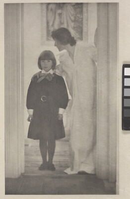 A soft-focus, black-and-white photograph of a White woman in a long, white dress with her arm around the shoulders of a White girl wearing a dark dress standing in a door frame.