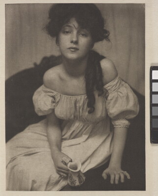A black-and-white portrait photograph of a seated young White woman dressed in white and leaning forward to gaze into the camera.