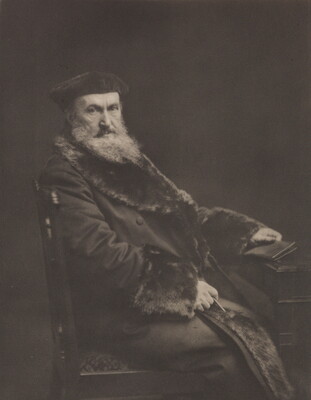 A black-and-white portrait photograph of a seated older White man with a long beard, wearing a beret and a long, fur-lined coat, holding a book in one hand.