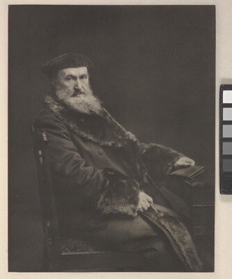 A black-and-white portrait photograph of a seated older White man with a long beard, wearing a beret and a long, fur-lined coat, holding a book in one hand.