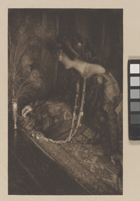 A sepia-toned photograph of a seated White woman wearing a long, patterned dress and a long, beaded necklace leaning toward a vase filled with peacock feathers.