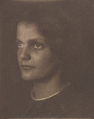A sepia-toned portrait photograph of a young White woman, dark hair blending into the background, gazing to the side.