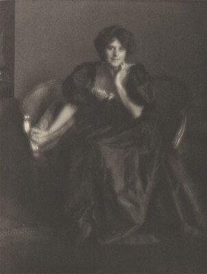 A black-and-white portrait photograph of a White woman, hair in an updo, wearing a long formal dress with puffy sleeves seated on a small sofa.