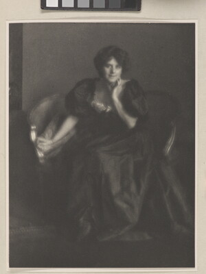 A black-and-white portrait photograph of a White woman, hair in an updo, wearing a long formal dress with puffy sleeves seated on a small sofa.