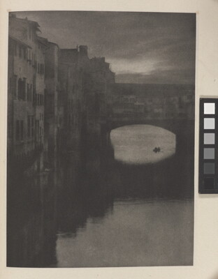 A sepia-toned photograph of a wall of tall buildings lining a river and an enclosed arched bridge stretching over the water.