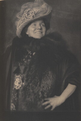 A black-and-white photograph of a smiling White woman wearing a hat decorated with pearls and veiling, a fur stole, and a dark robe or jacket, one hand on her hip.