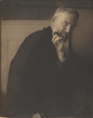 A soft-focus sepia-toned photograph of an older White man with short hair and a mustache, chin resting on one hand.