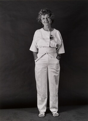 A black-and-white portrait photograph of a standing older White woman with short curly hair, hands in her pockets, wearing a white top, pants and sandals.
