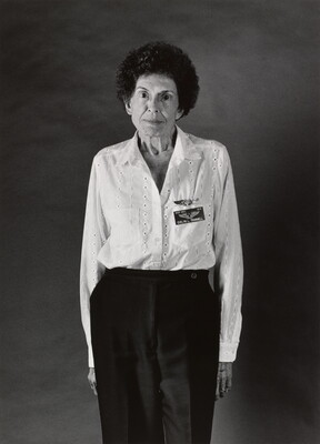 A black-and-white photograph of an elderly White woman with short, curly hair wearing a white blouse with a name tag, standing with her arms to her side.