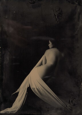 A black-and-white photograph of a seated nude White woman lightly draped in fabric with her back to the camera.