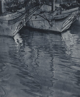 A black-and-white photograph of the ends of two docked sailboats floating on rippling water.