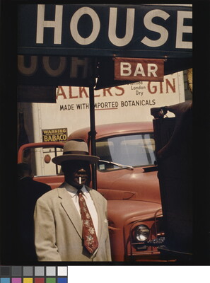 A color photograph of a Black man in a beige hat and suit with a cigarette in his mouth in front of a red car and under a sign that says, "House Bar."