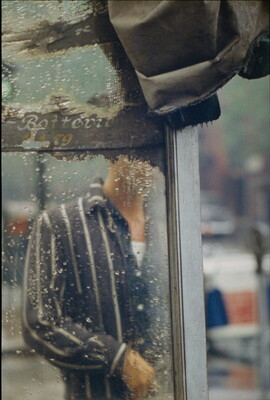A color photograph of a man's torso through the rain-splatted glass of what looks like a bus shelter.