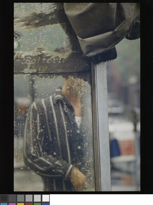A color photograph of a man's torso through the rain-splatted glass of what looks like a bus shelter.