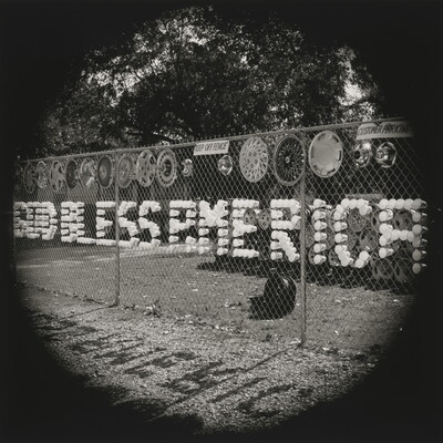 A black-and-white photograph of plastic cups arranged in a chain-link fence to spell out "God Bless America."