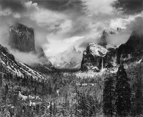 A black-and-white landscape photograph of trees and snowy mountains partially covered in clouds and fog.