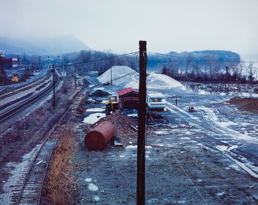 A color photograph of a railyard in winter with rusty barrels, piles of sand, machinery, and patches of snow scattered around with trees and a foggy mountain in the distance.