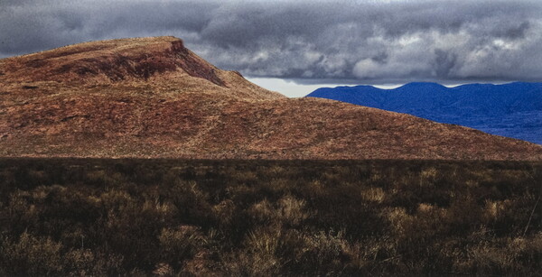 A color photograph of a desert landscape with a dry, brown hill in the foreground and blue mountains in the background under a sky of gray clouds.