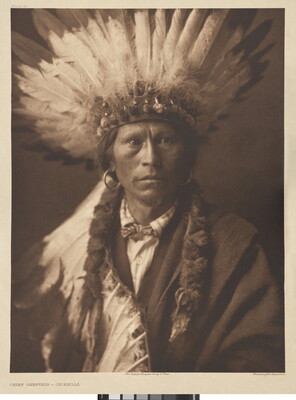 A sepia-toned photograph of a Native American man posed in a suit and feathered headdress.