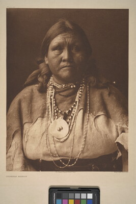 A sepia-toned portrait photograph of an older Indigenous woman with her hair bound on both sides of her head and wearing several beaded necklaces and traditional clothing.