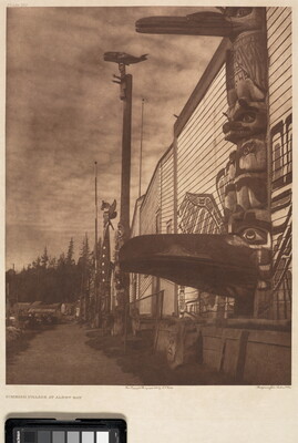 A sepia-toned photograph of several totem poles pressed next to a building with a mural painted on the side.