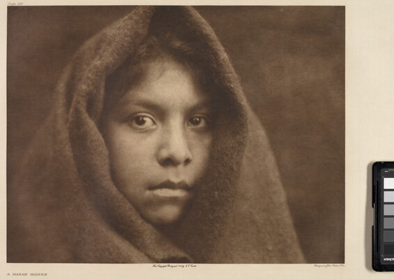 A sepia-toned portrait photograph of a young Indigenous girl wearing a cloth over her head, exposing only her face and hair above her forehead.