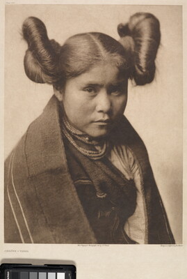 A sepia-toned portrait photograph of a young Indigenous woman with her hair split into two large buns on both sides of her head wearing traditional clothes and necklaces.