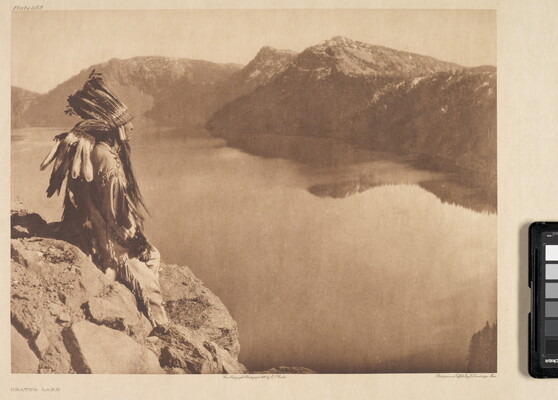 A sepia-toned photograph of an Indigenous man in traditional clothing and a feathered headdress sitting on a rock high above a lake at the base of mountains.