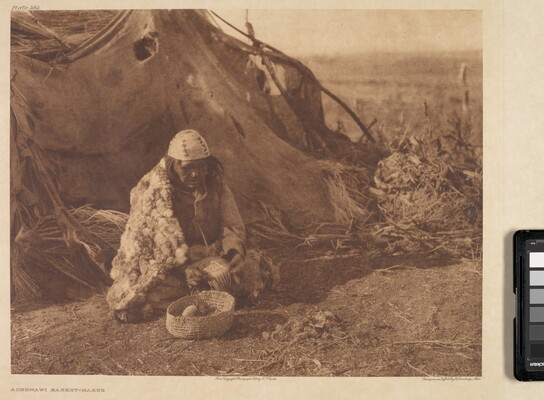 A sepia-toned photograph of an Indigenous person in traditional dress sitting on the ground outside of a hut with a woven basket in front of them.