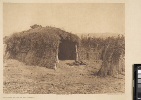 A sepia-toned photograph of a hut made of grass, brush, and wood on a dirt clearing.