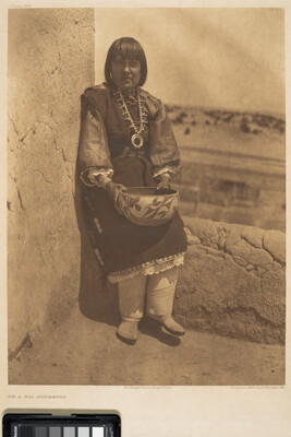 A sepia-toned photograph of an Indigenous woman in traditional dress holding a bowl seated on the ledge of a clay-style building.