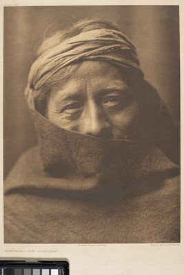 A sepia-toned portrait photograph of an older Indigenous person wearing headband with their face partially covered by what looks like a blanket.