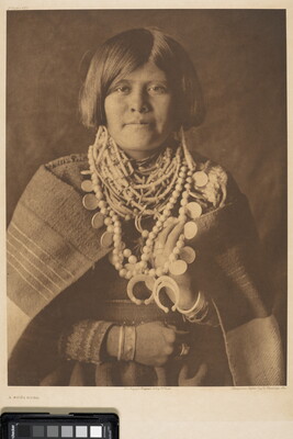 A sepia-toned portrait photograph of a young Indigenous person with short, bobbed hair, wearing many beaded necklaces, and a blanket wrapped around her shoulders.