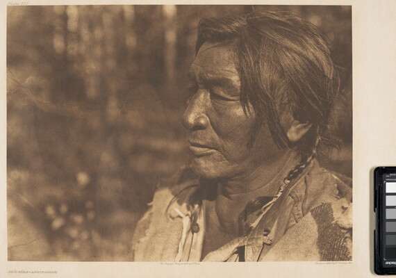 A sepia-toned photograph of the close up of a middle-aged Native American man's face, in profile, with two braids.