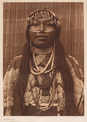 A sepia-toned portrait photograph of an Indigenous woman wearing a beaded hair covering over her braids, a long quill through the septum of her nose, beaded necklaces, and an elaborately beaded tunic.