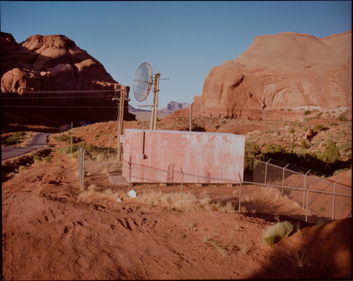 A color photograph of a small rectangular structure with a satellite behind a chain-link fence in the middle of a desert with many red rock formations.