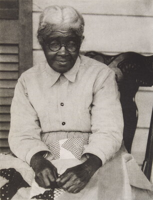 A black-and-white photograph of a seated, elderly Black woman holding a quilt on her lap.